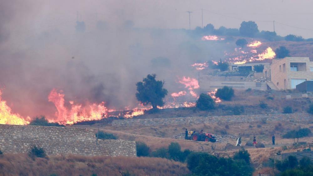 A fire burns through low-lying vegetation across an arid slope towards a village as people watch from nearby.