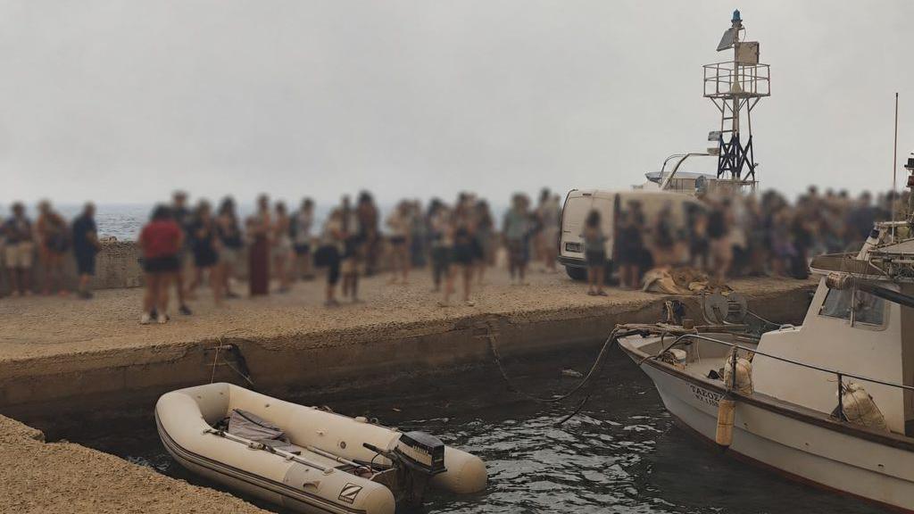 Blurred-out people stand on a marina pier while waiting to be evacuated in Agios Vasileios in Greece.