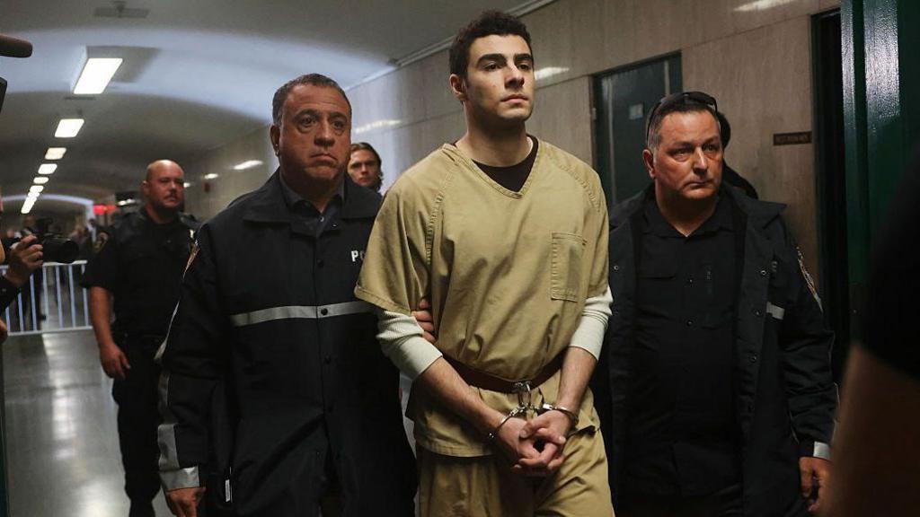Mangione walking, in jail outfit and handcuffs, led by two guards