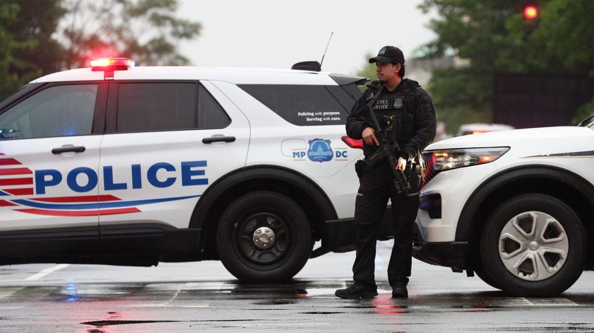 A police officer with a gun stands in front of two white marked police cars