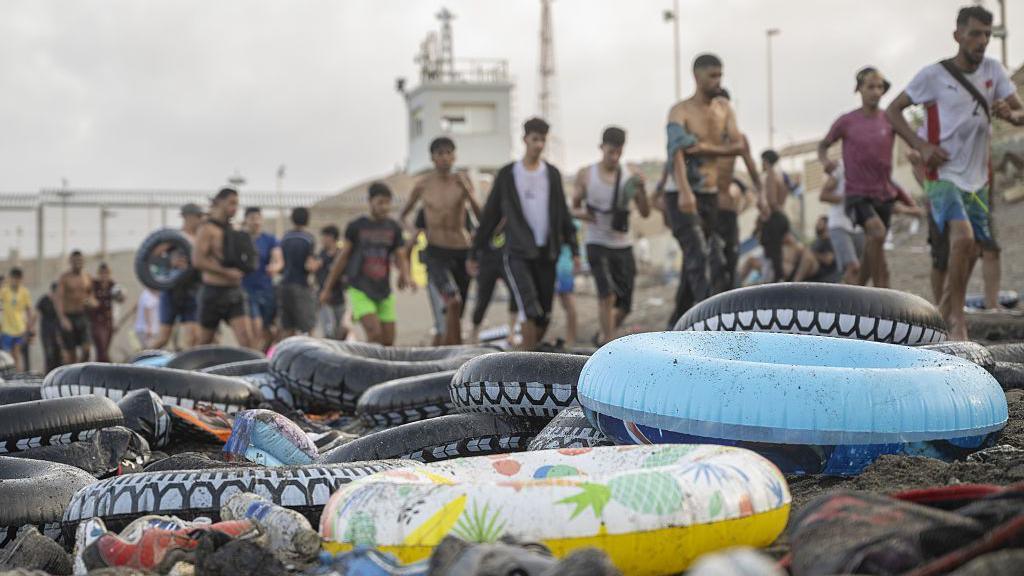 Rubber flotation rings sit on a beach in front of a line of arriving migrants in Ceuta.