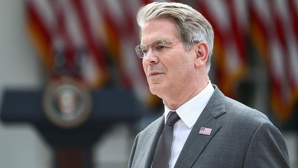 US Treasury Secretary Scott Bessent's head and shoulders in profile. He has grey hair and glasses. He is wearing a grey suit with a US flag lapel pin, a silver tie, and a white shirt. Behind him in soft focus are US flags and a podium.