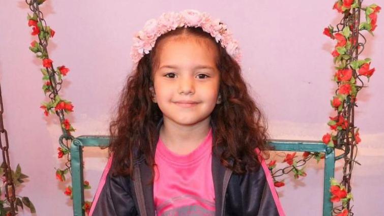 Hind Rajab smiles at the camera. She has long, brown wavy hair and is wearing a pink flower crown on her head. She also has a pink T-shirt and black zip-up on. She is sitting on a chair adorned with garlands of red flowers.