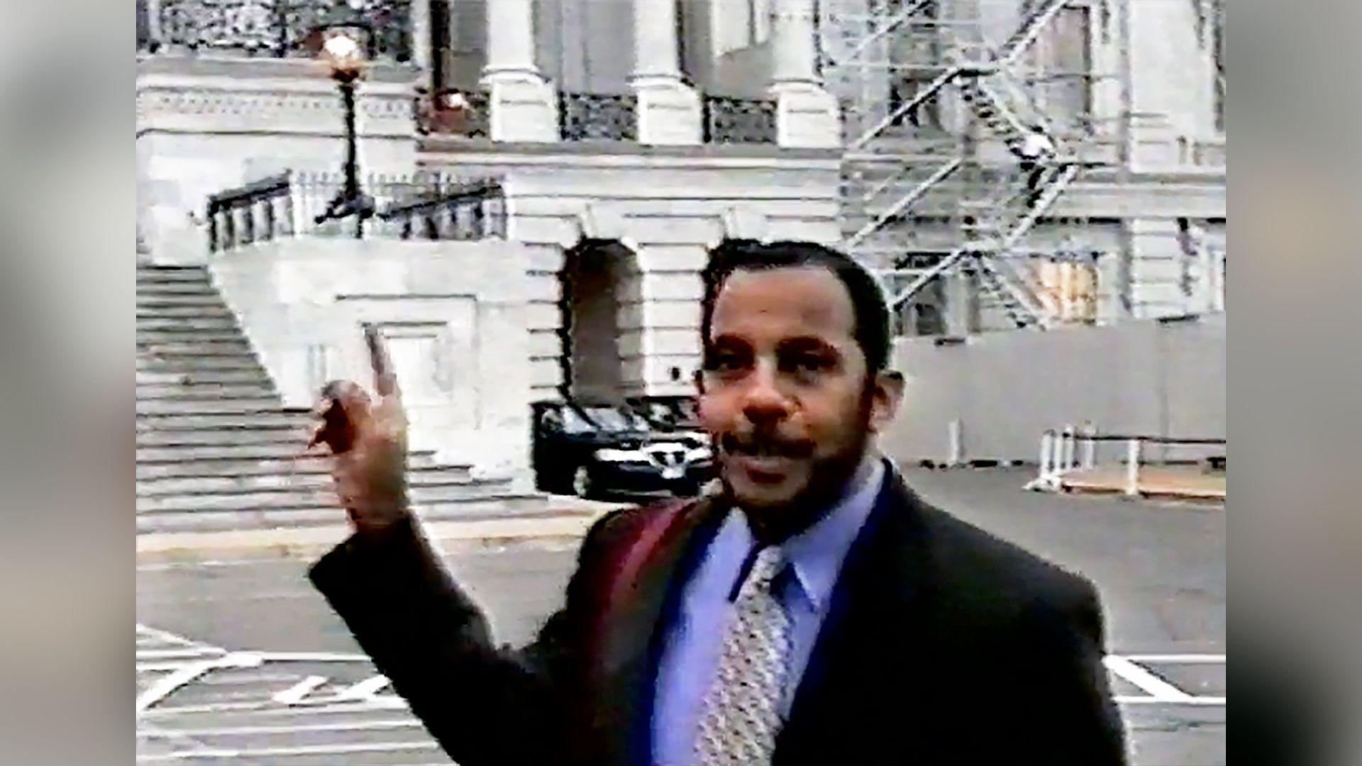 A screenshot of Bayoumi, wearing a suit with a purple shirt, standing outside the Capitol. He is pointing towards the building.