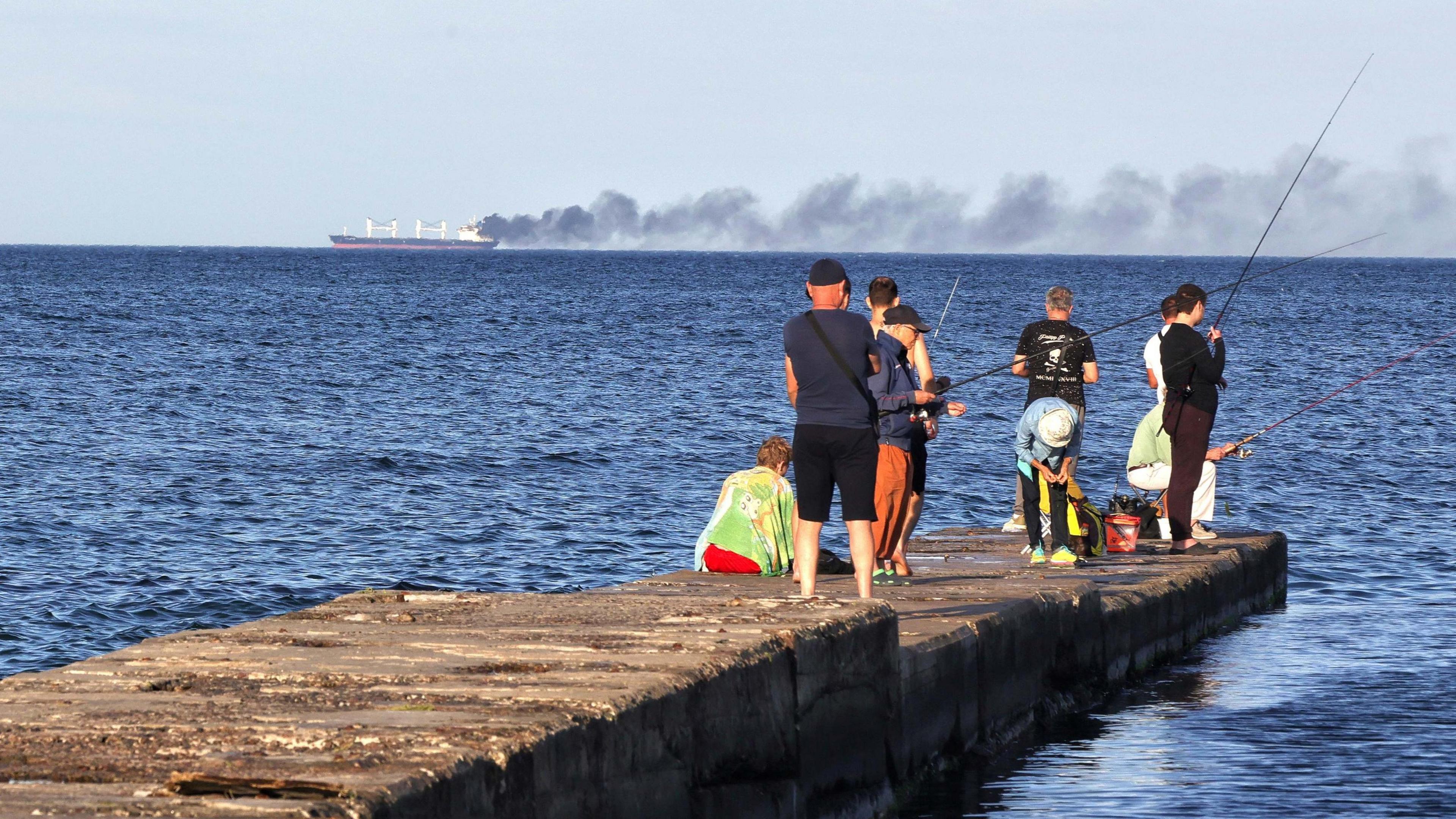 Men fish on a pier as black smoke rises from a vessel damaged by a Russian attack in the Black Sea, Odesa region, Ukraine, July 28, 2026