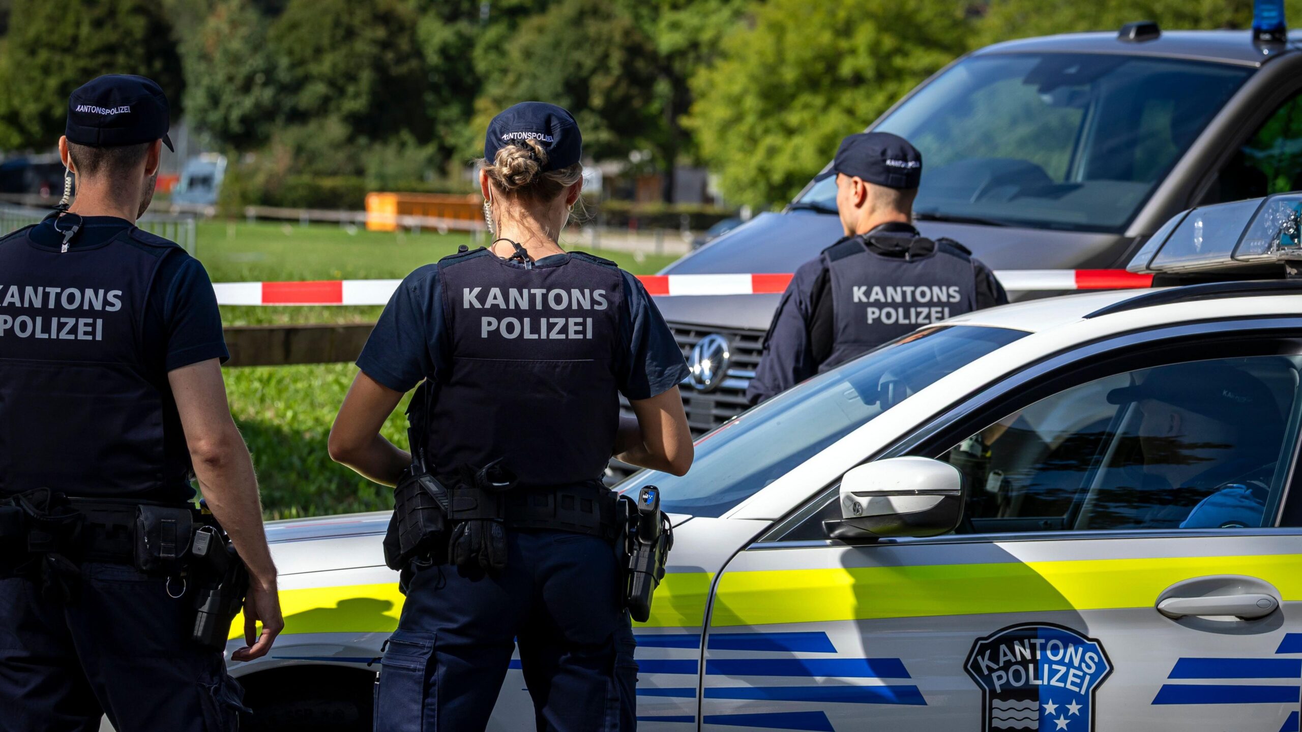 Three police officers in uniform, including a bulletproof vest reading Kantons Polizei at the back, stand watching a field cordoned off by white and red tape. A fourth officer sits inside a police car parked next to his colleagues