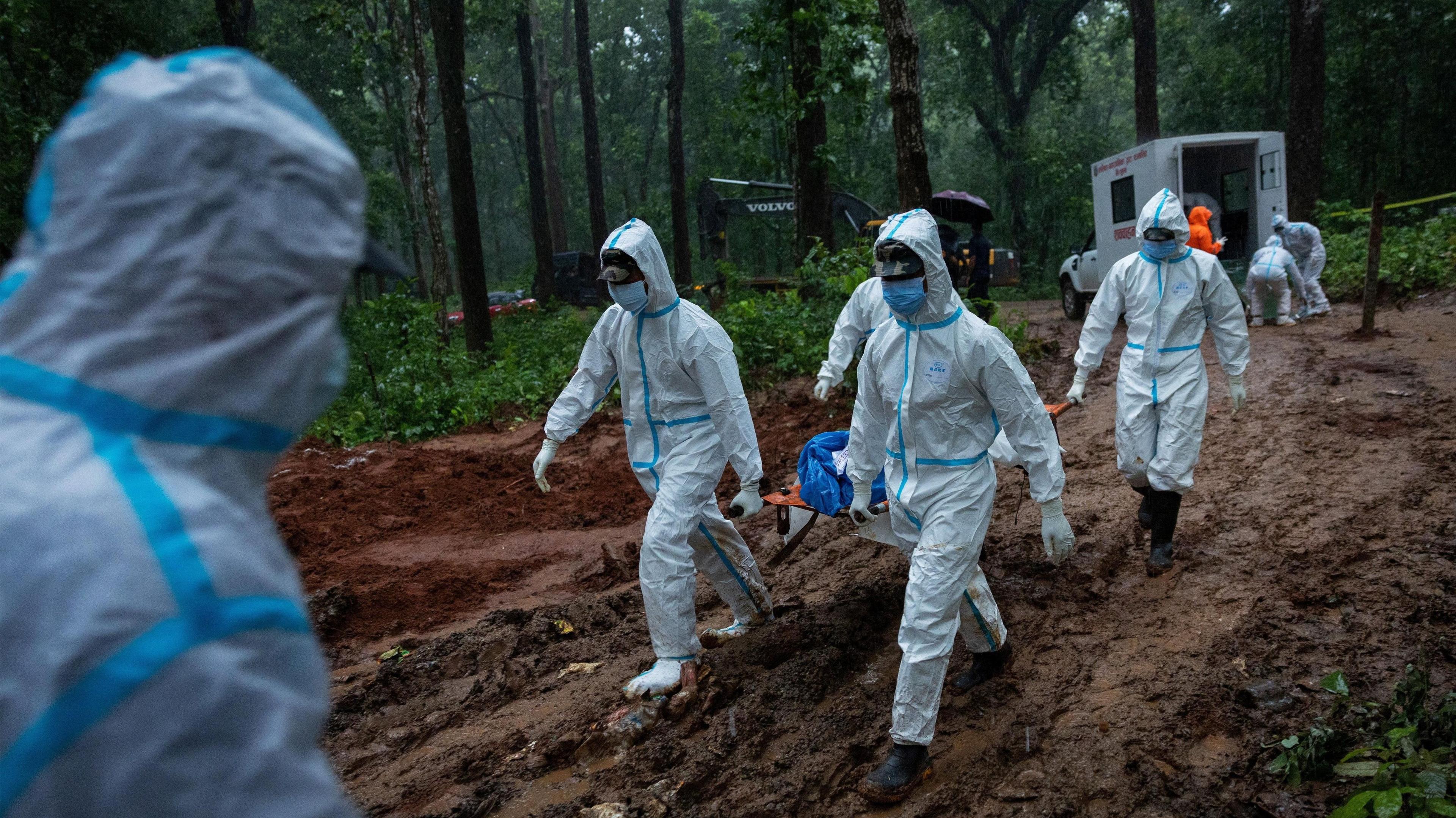 People in white PPE carry a stretcher with a dead body on it in a forest