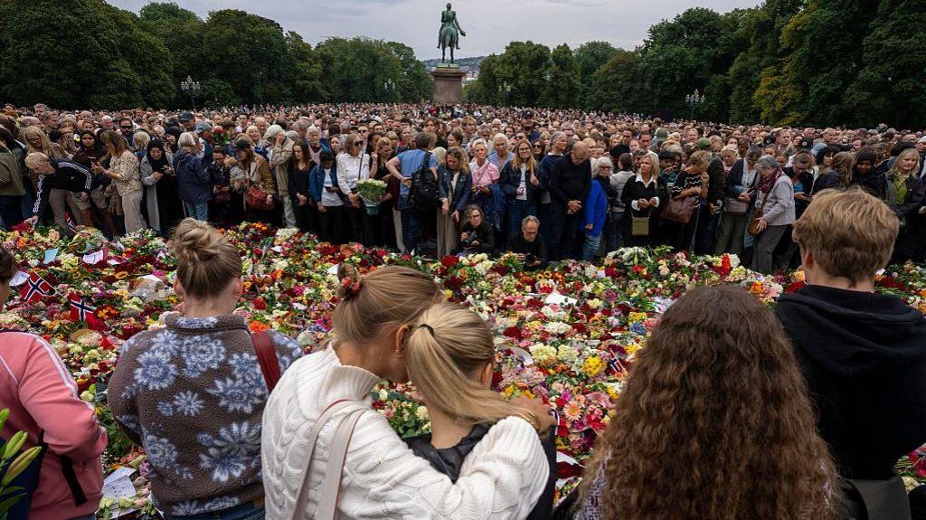 People stand in a crowd, two women hugging each other, against a line of flowers and flags on the ground, a statue behind them at the royal palace in Oslo.