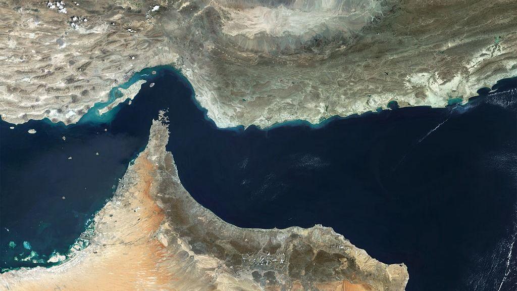 A satellite view of the Strait of Hormuz, with a thin stream of dark blue water flowing around a sharp point of arid land jutting into it.