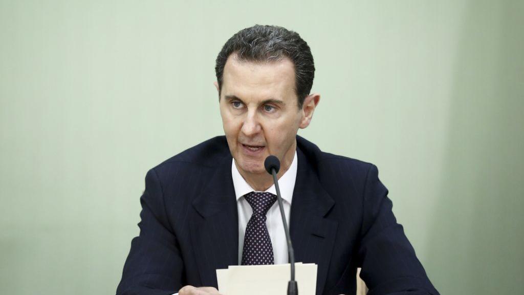 Bashar al-Assad pictured in 2023 speaking into a microphone and holding some papers. He is wearing a dark suit, white shirt and tie.