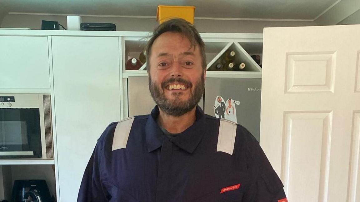Zach standing in a kitchen wearing a blue military boiler suit and smiling at the camera. He has a short beard and brown hair.