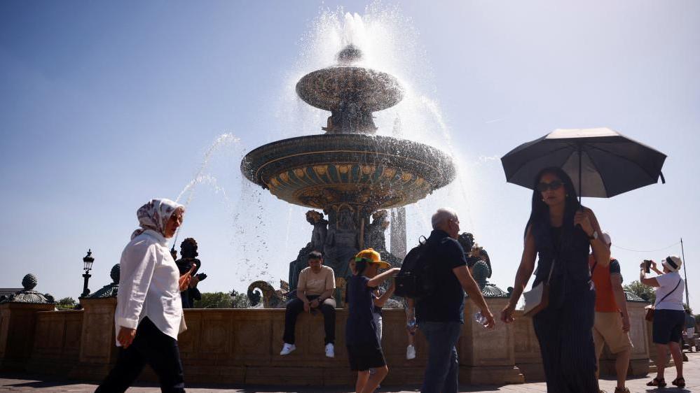 People walk around a fountain, with blue sky and bright sunshine in the background. One woman is holding an umbrella for shade; other people are wearing sunhats and carrying bottles of water.