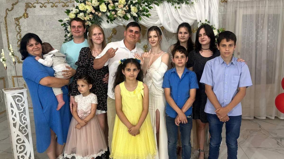 Twelve family members smile at the camera while what appears to be a bride and groom hold up their traditional wedding ring fingers