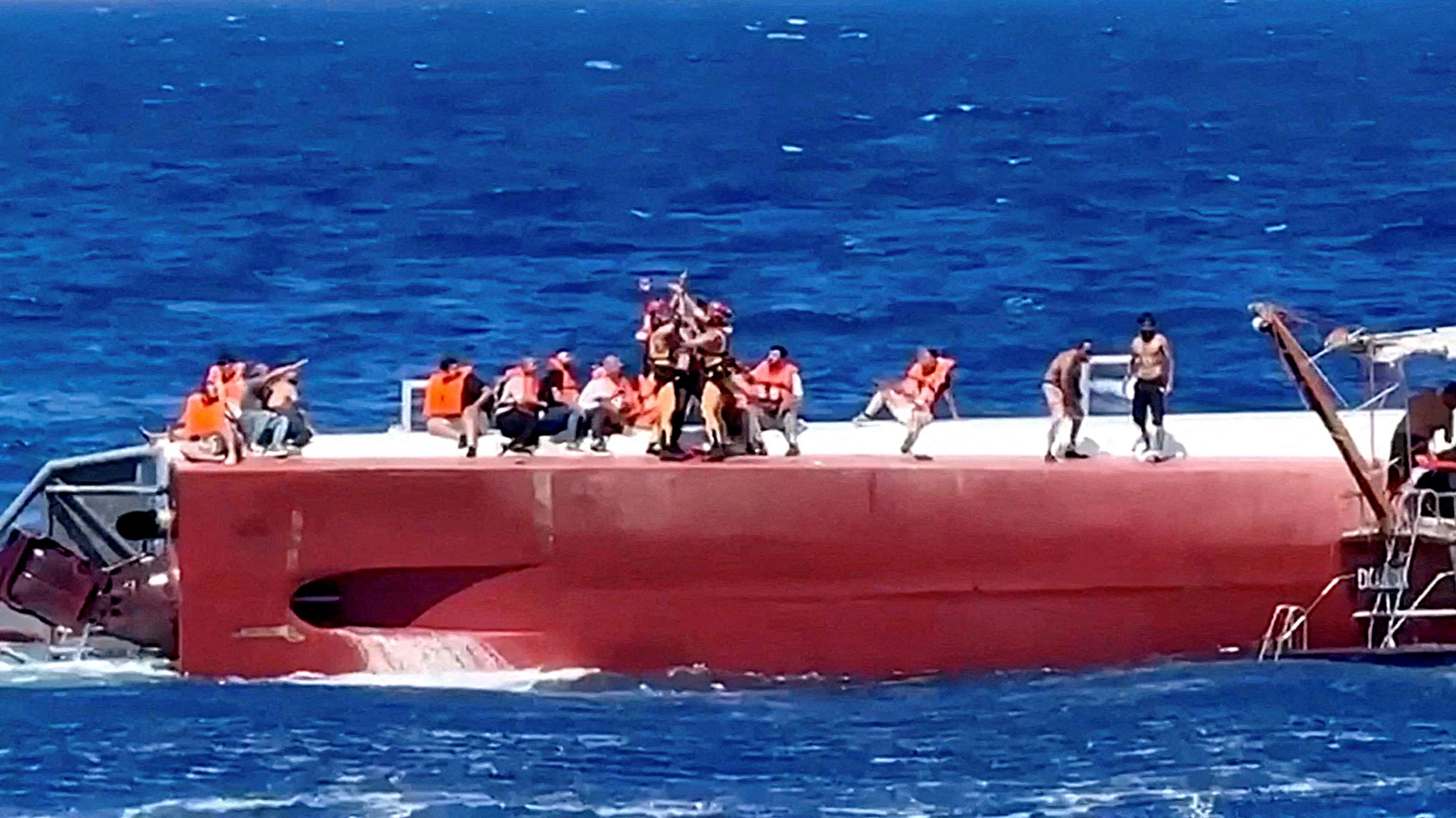 People wearing life jackets sit atop a capsized ferry as rescuers airlift one to safety.