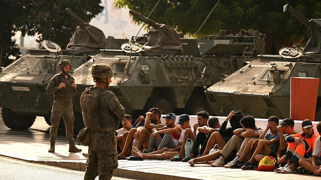 Military personnel stand near armored vehicles while a group of seated people wait on a curb beside a road, under trees in daylight