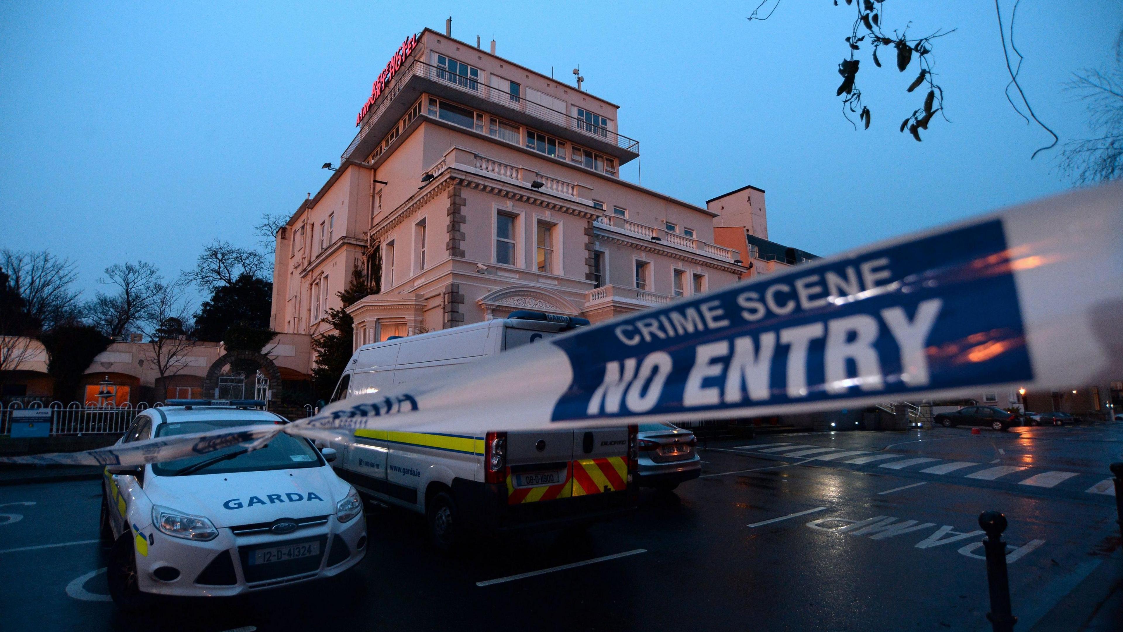 A garda (Irish police) cordon outside the Regency Hotel in Dublin in 2016. A white and blue liveried garda van and car are parked outside a four-storey hotel at dusk on a wet evening. A white a blue cordon tape stretches across an entrance to the hotel car park.