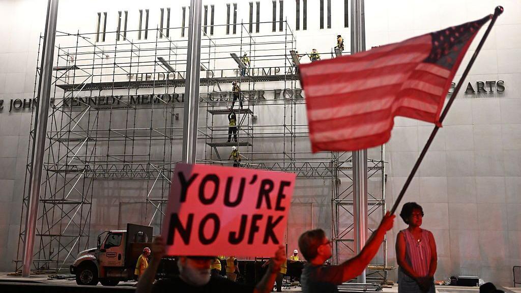 people gathered in front of Kennedy Center building, one holding an American flag and another holding a sign that says "you're no JFK"