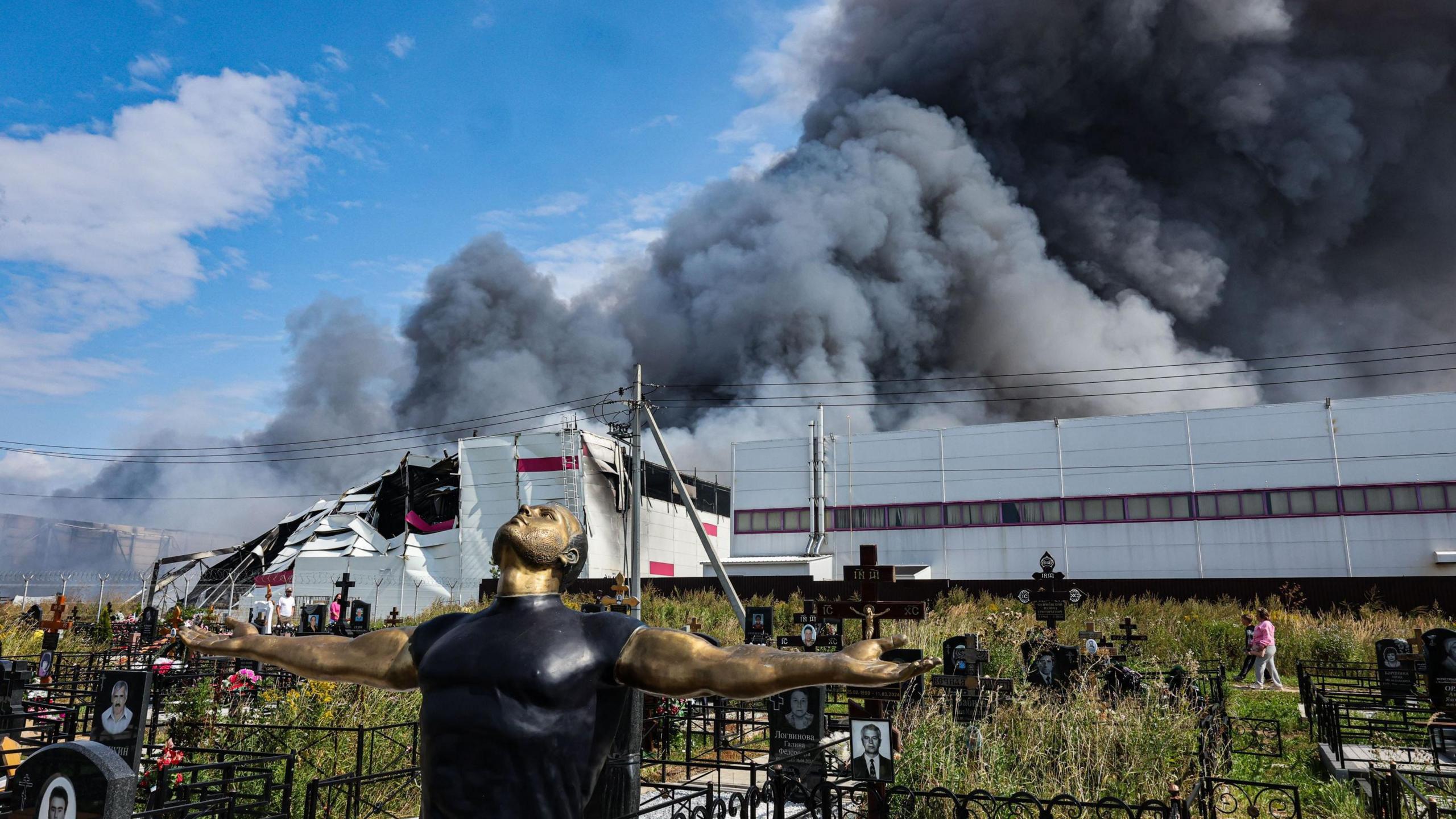 People walk among graves at a cemetery as smoke rises above a Wildberries facility (back) following a reported Ukrainian drone strike at the Koledino Industrial Park near Podolsk, Moscow Region, Russia, 16 August 2026