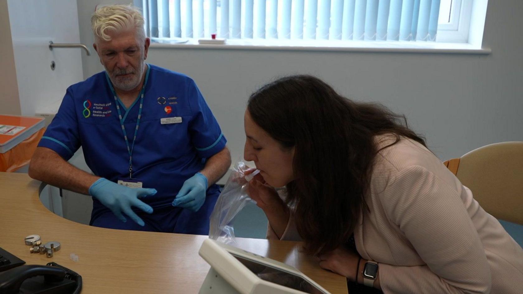 A woman with long dark hair and wearing a light pink jacket leans forward towards a desk. She is blowing into a tube on a clear plastic bag which is attached to a small white machine with a built in monitor. To her left is a nurse watching her progress. He is wearing royal blue scrubs and blue latex gloves and has white hair and a short white beard.