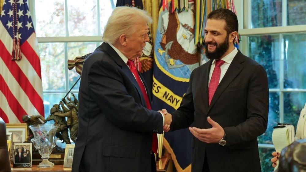 Two men in dark suits and red ties smile and shake hands in front of windows and American flags