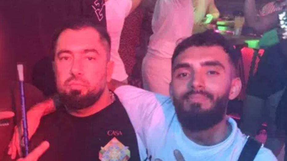 Two bearded men sitting in what appears to be a nightclub with people dancing behind them