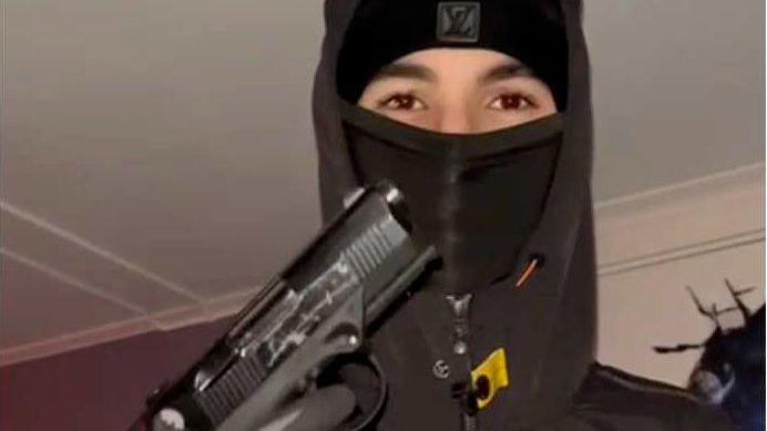 A teenager wearing a black balaclava and hoodie holding up a handgun, with his gloved hand on the trigger