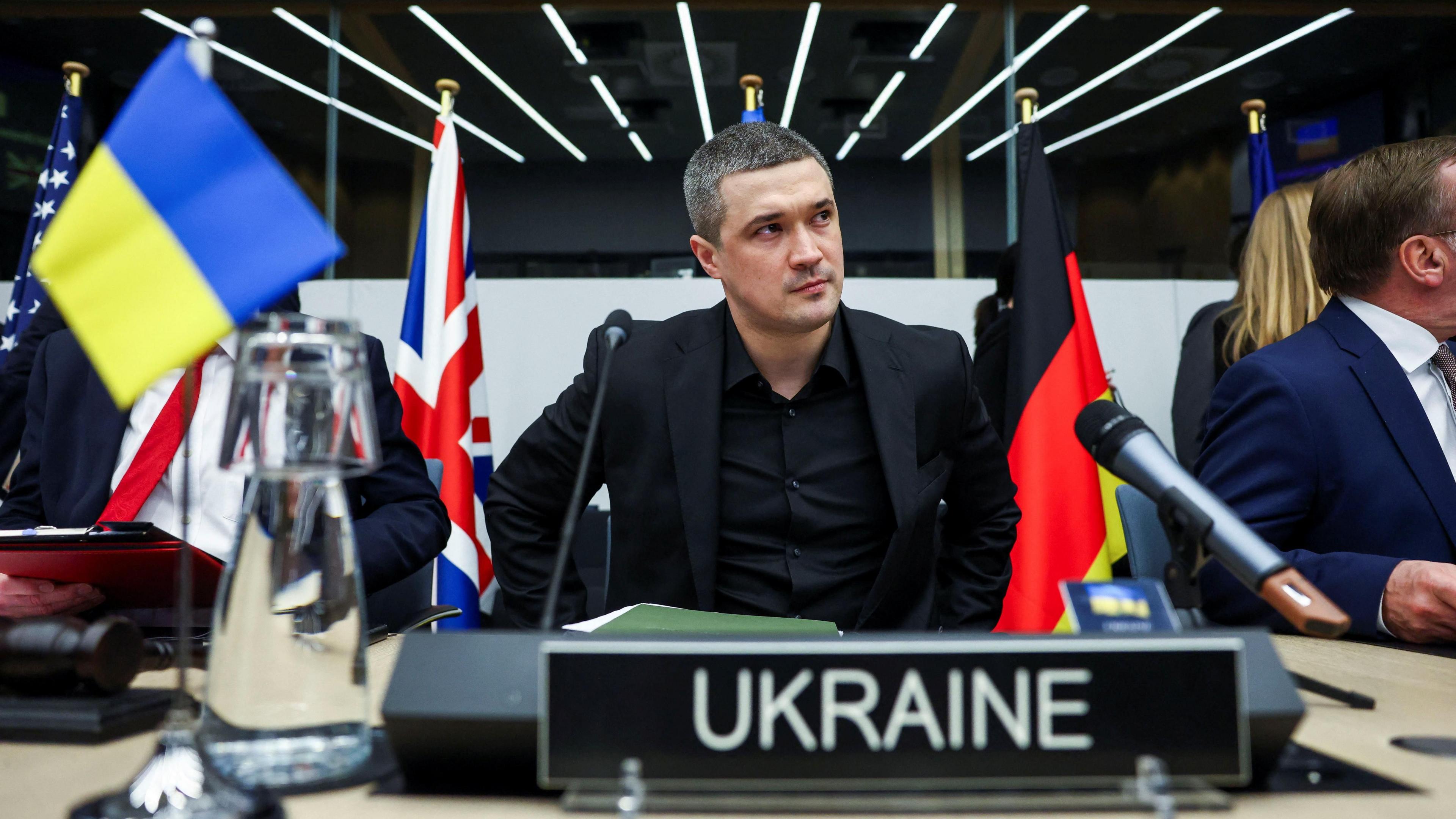 A man in a black suit and shirt sits between other people at a desk with countries' flags in the background and a table sign at the front that reads Ukraine next to a Ukrainian flag.