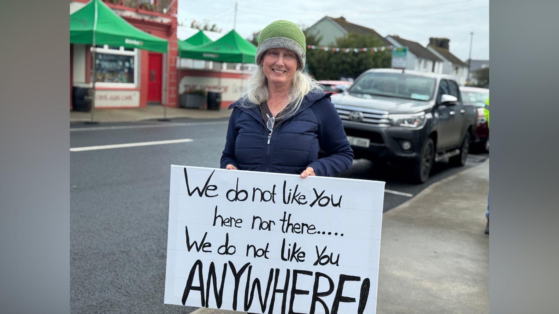 Jessie Phillips, blonde hair, green beanie hat, with her placard saying 'We do not like you here nor there.....we do not like you anywhere!' She is wearing a navy outfit.