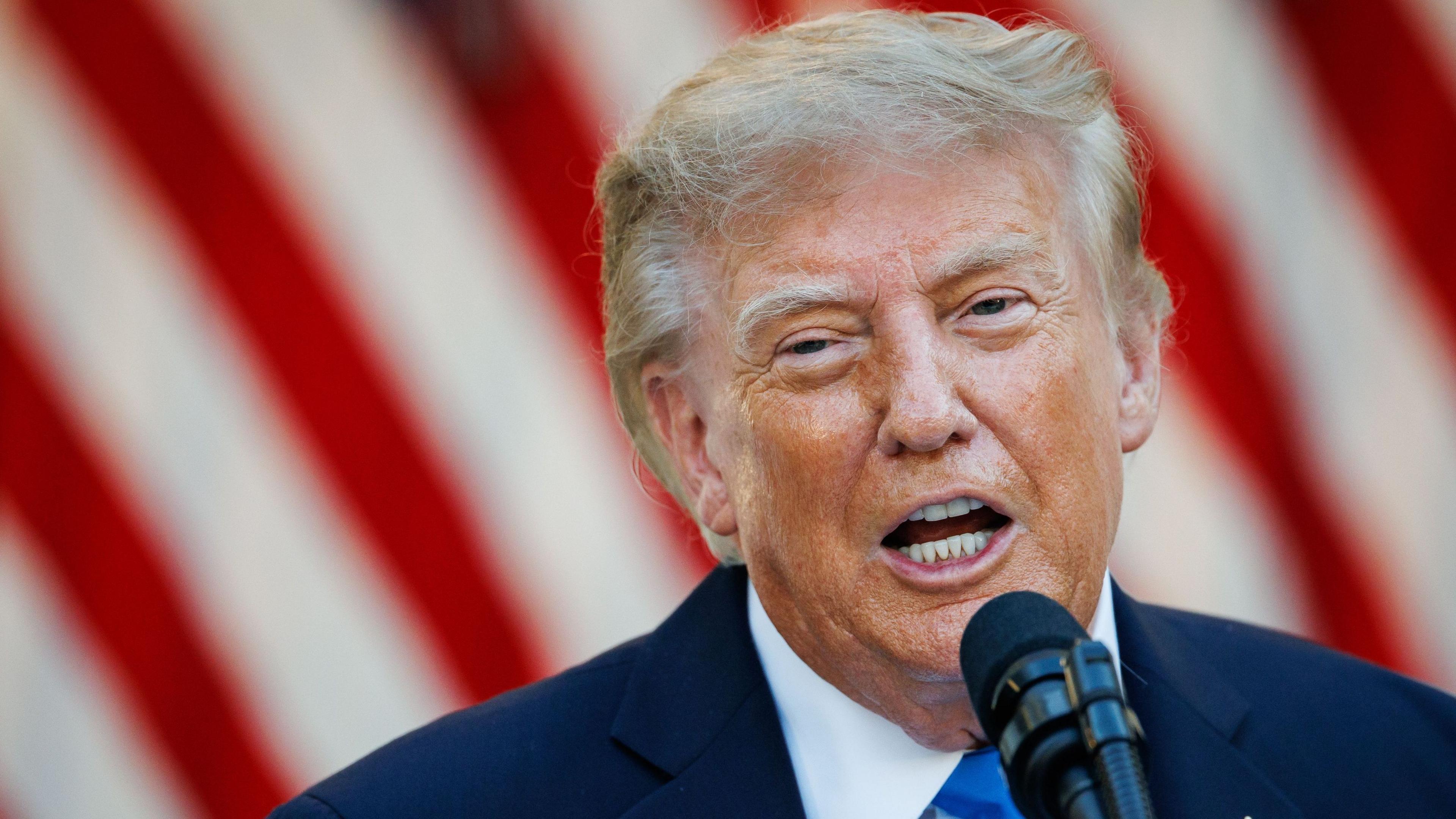 Closeup of Donald Trump speaking into a mic. Behind him are the red and white stripes of the American flag.