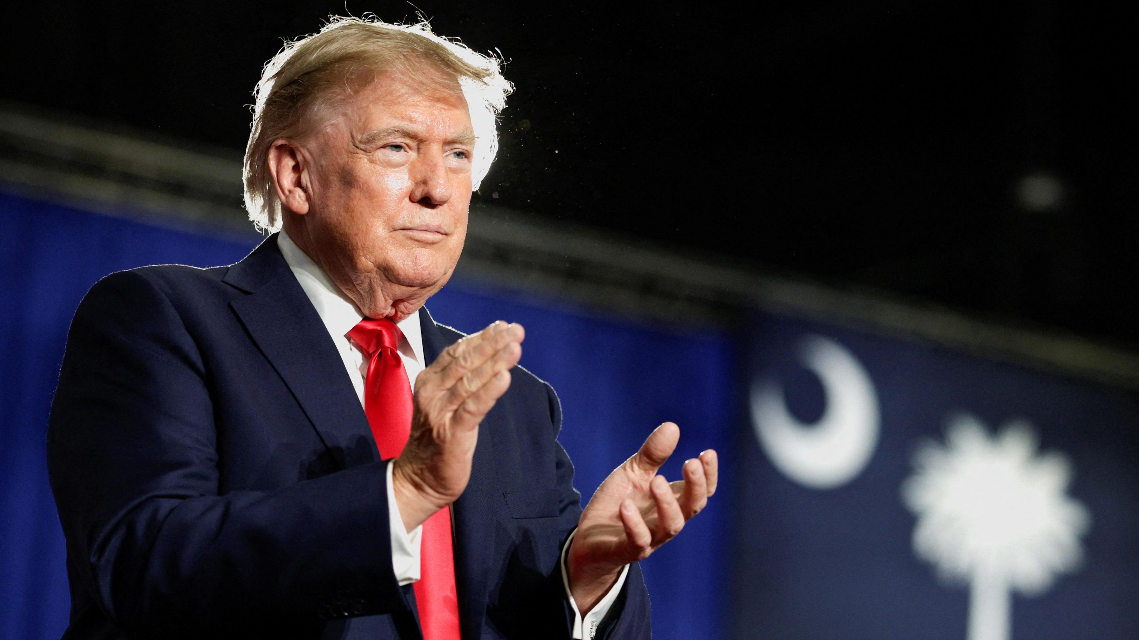 US President Donald Trump standing on a stage dressed in a navy suit and red tie clapping to an audience not in view.
