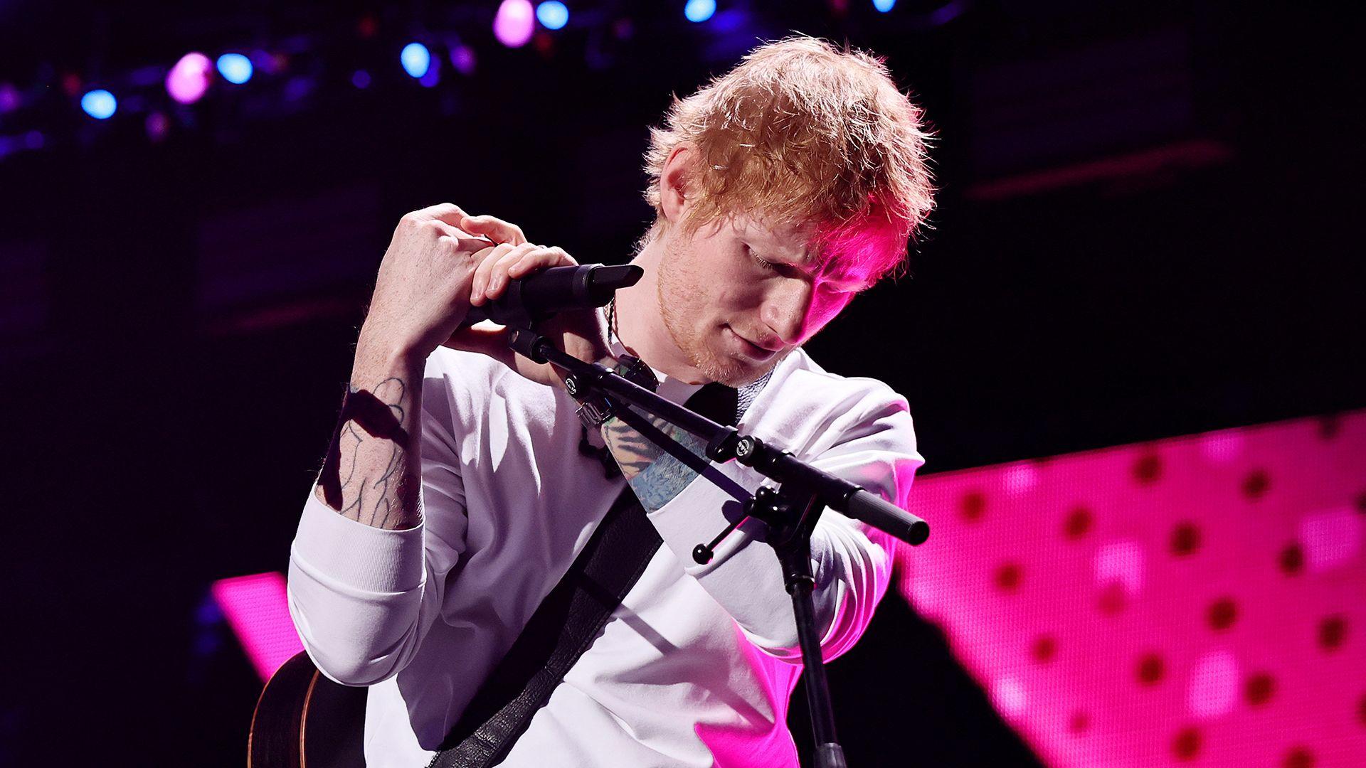 Ed Sheeran looks down at stage while holding a microphone in a mic stand against a pink and black background as he performs onstage in New York in December.