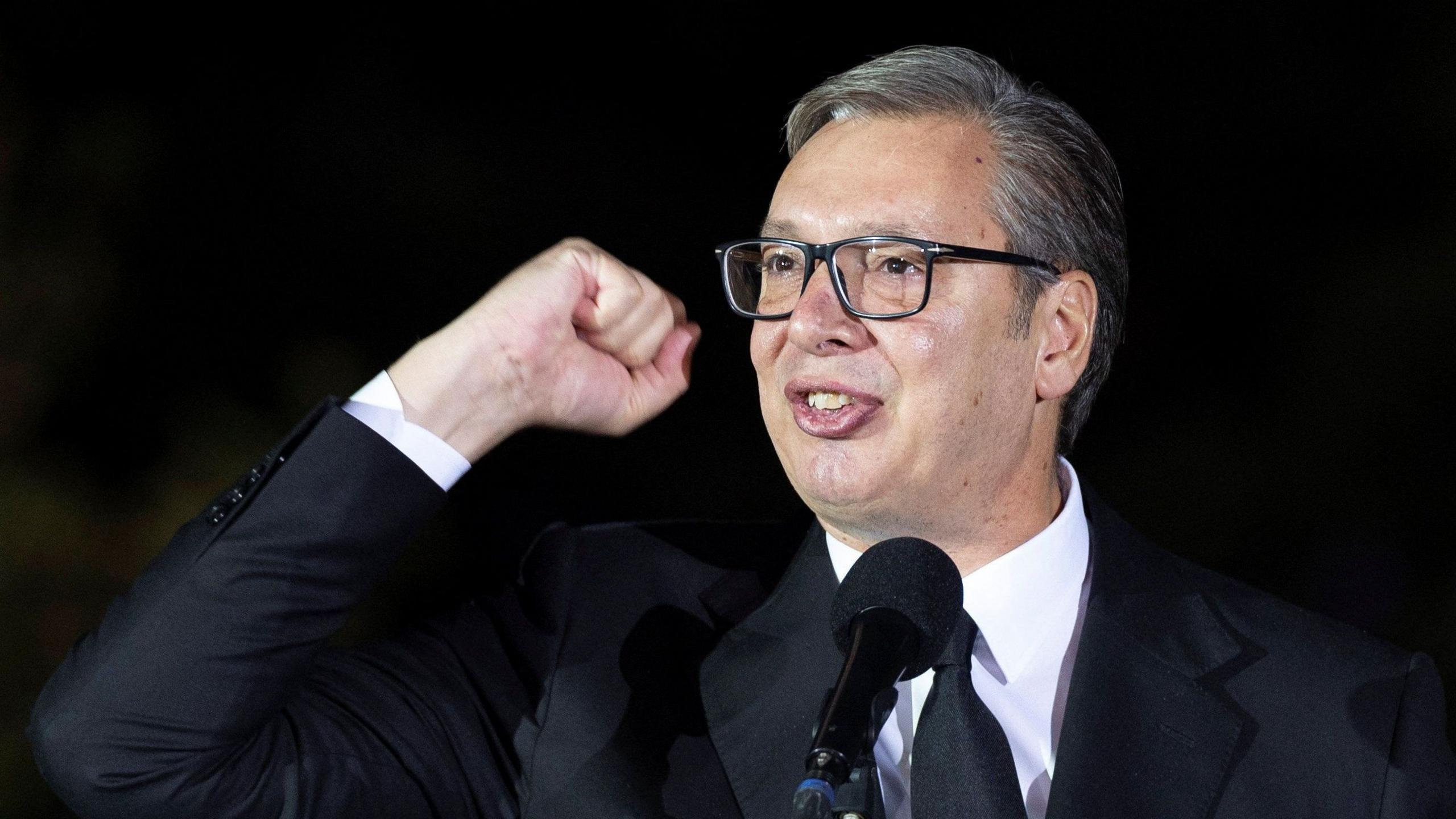 A head shot of Aleksandar Vucic looking off-camera giving a speech while waving his fist in the air. He is wearing a black suit and tie with white shirt.