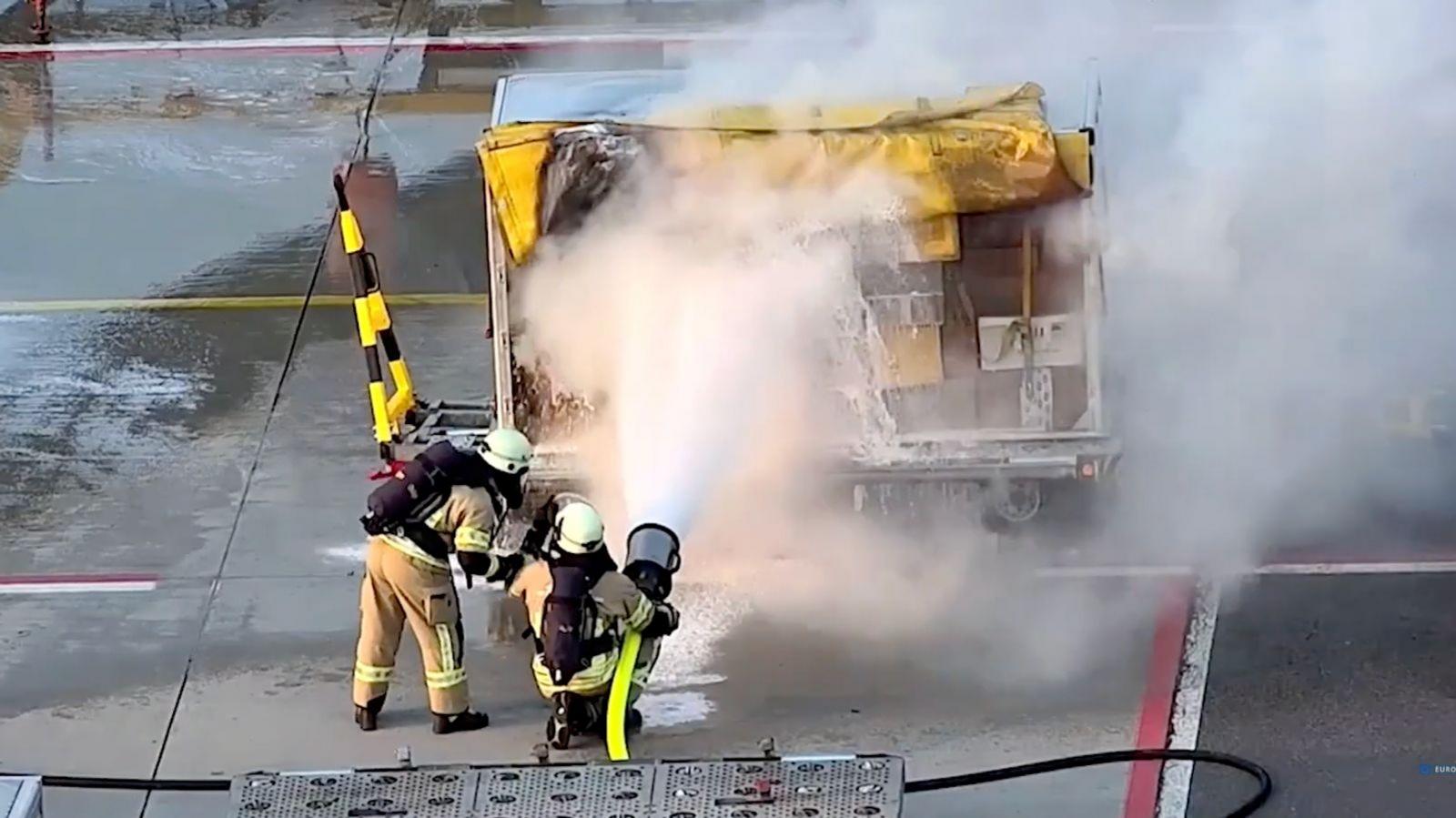 Two fire officers are spraying foam on an airport cargo trolley. Smoke is spreading from the trolly.