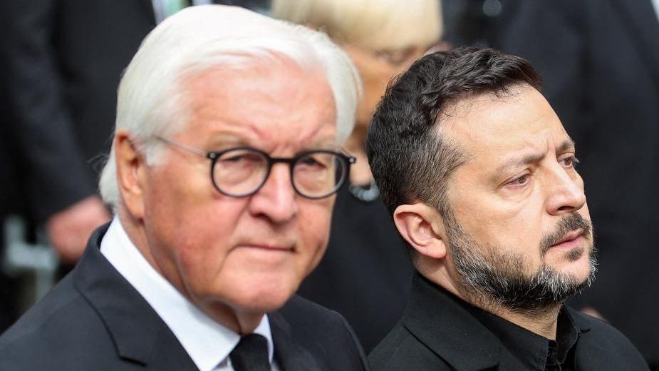 Frank-Walter Steinmeier, who has grey hair and wears glasses, looks on while wearing a suit as he walks next to Zelensky, who has short brown hair and wears a black shirt with black suit jacket