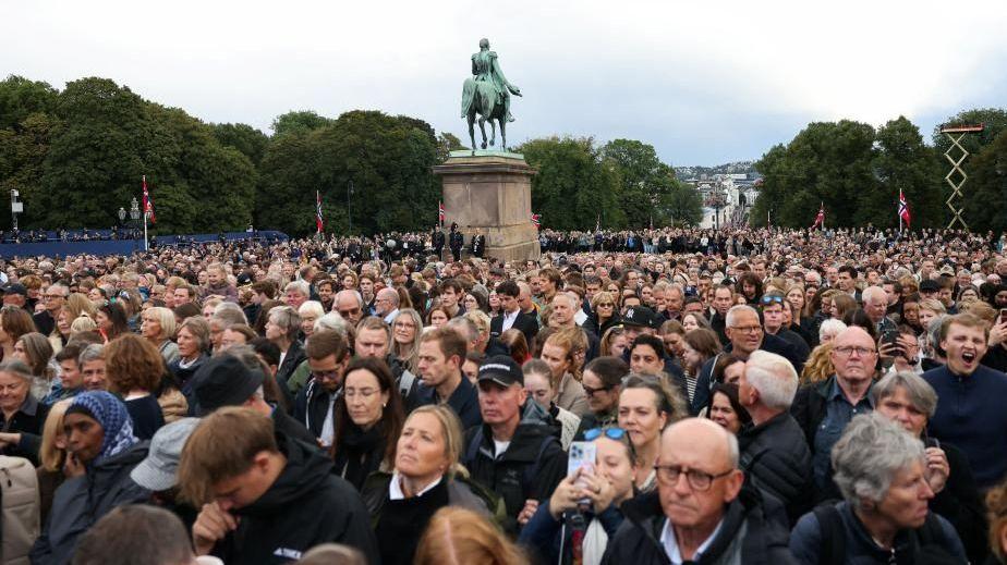 Hundreds of people, many with solemn faces, can be seen gathered in the square outside of the royal palace on Wednesday. A statue and trees are seen in the background