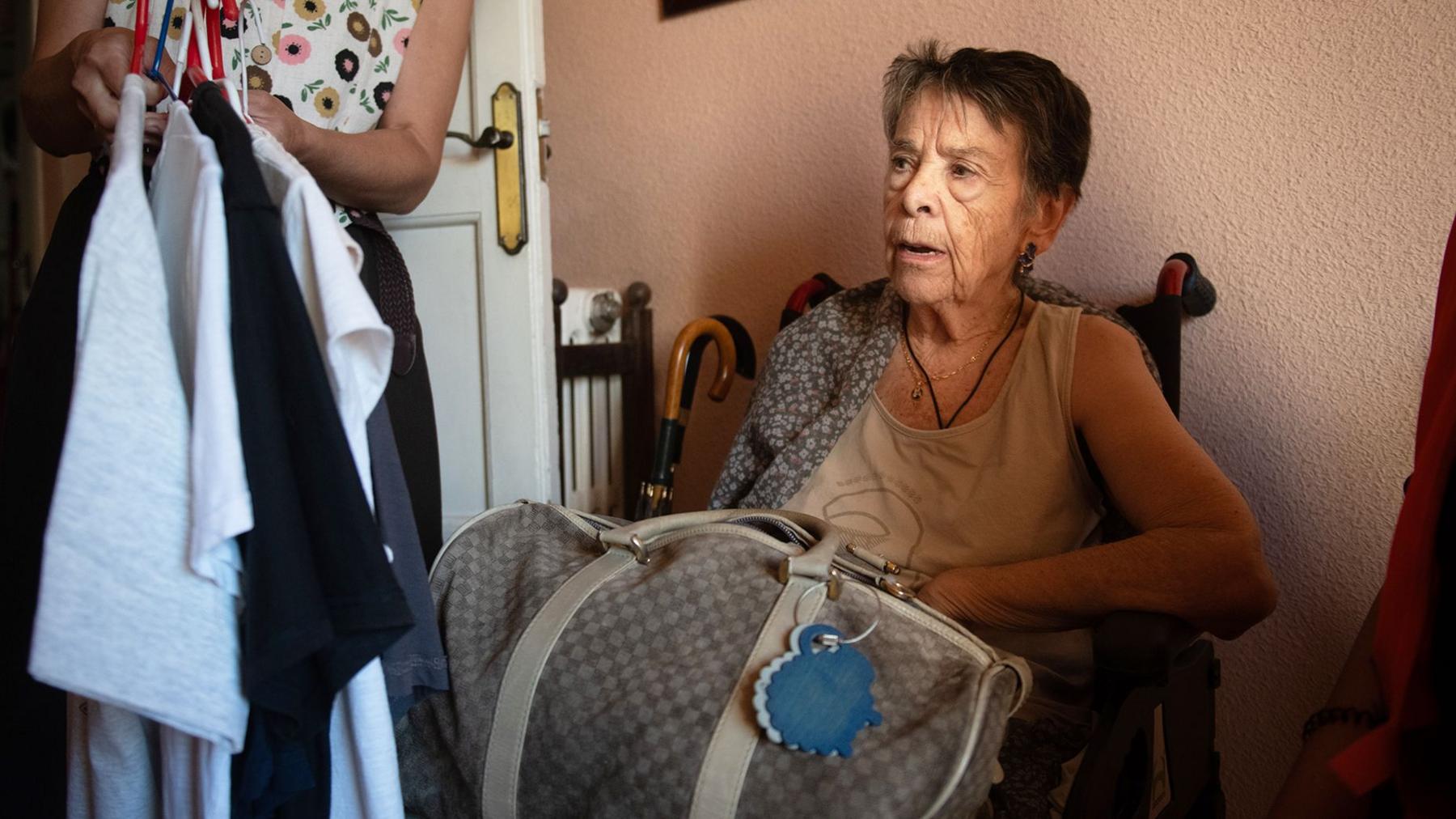 Maricarmen, a frail elderly woman, packs a bag with her belongings as she sits in a chair by the door of her flat