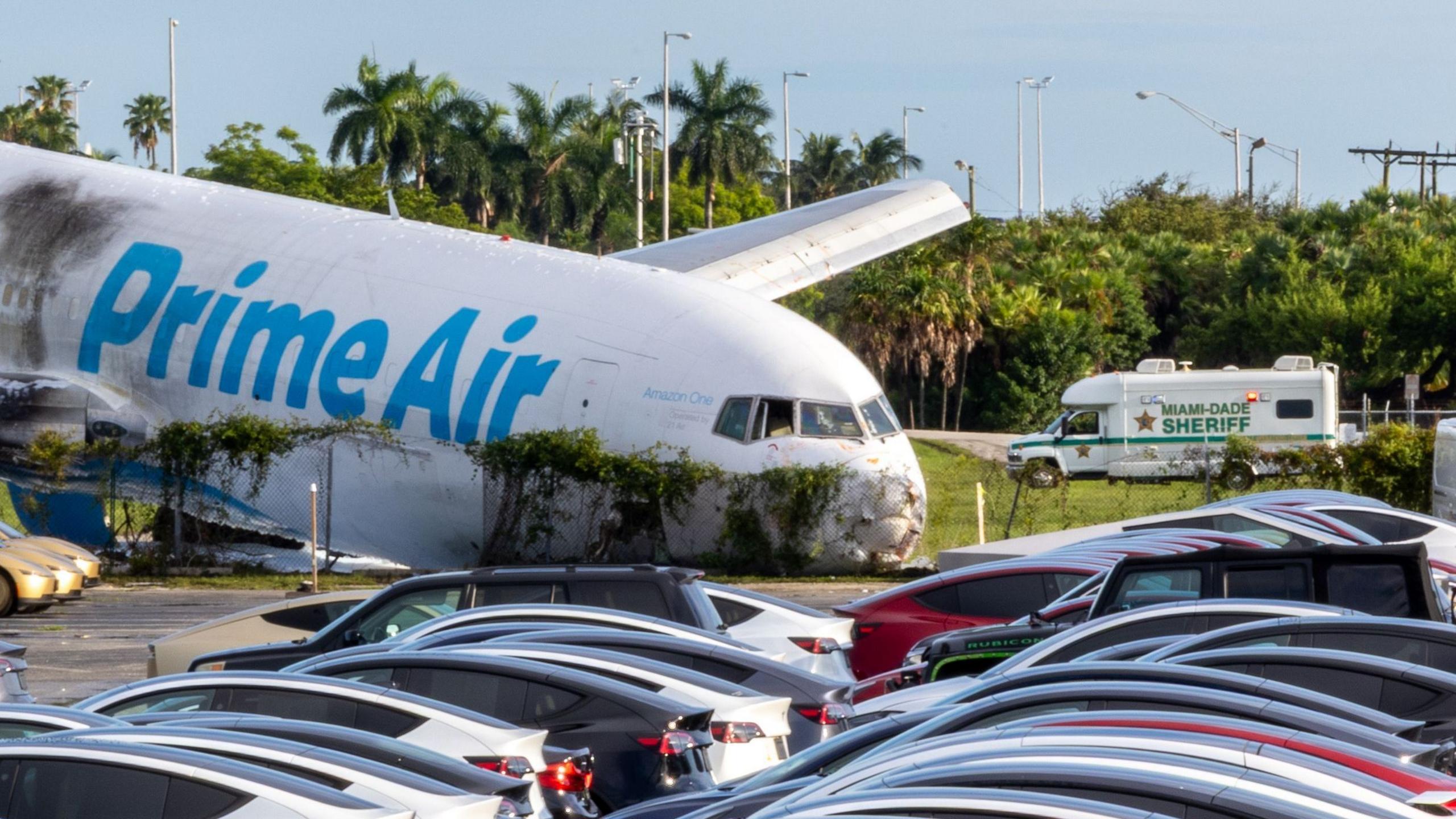 A Prime Air plane rests next to a car park after crashing. The nose of the plane is crumpled and there are black marks on the side of the plane.