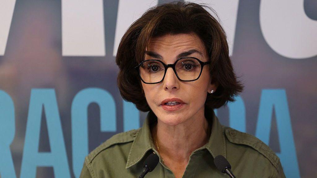 Rachida Dati speaks at a podium with two microphones, wearing dark-framed glasses and an olive-green shirt