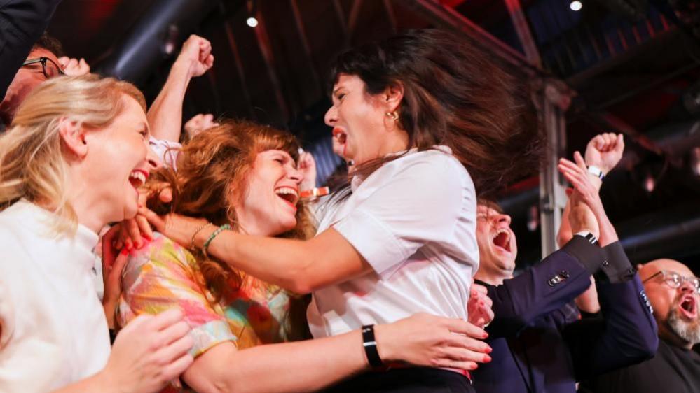 Elif, who has shoulder-length brown hair and wears a sleeveless white shirt, is seen cheering and hugging a woman and others look elated around them