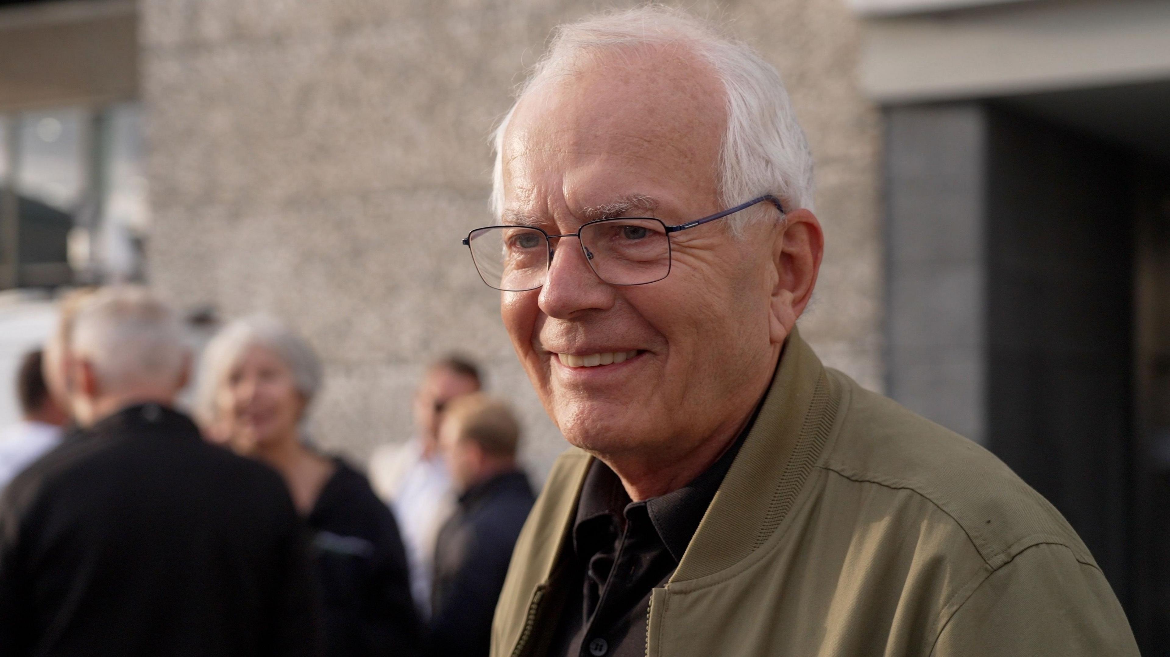 Reinhart. He is an older man with balding white hair. he has black rimmed glasses on and a beige unzipped jacket