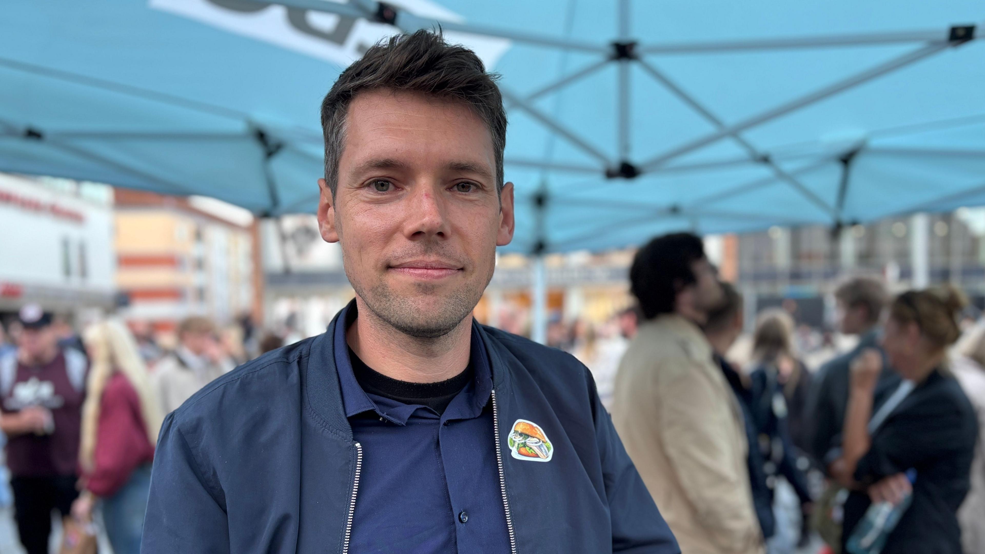 Rohloff close up. He is wearing a black shirt with a black unzipped jacket. There is a sticker of a sandwich on his left lapel. he is outdoors, standing beneath a canopy. There are people in the background.