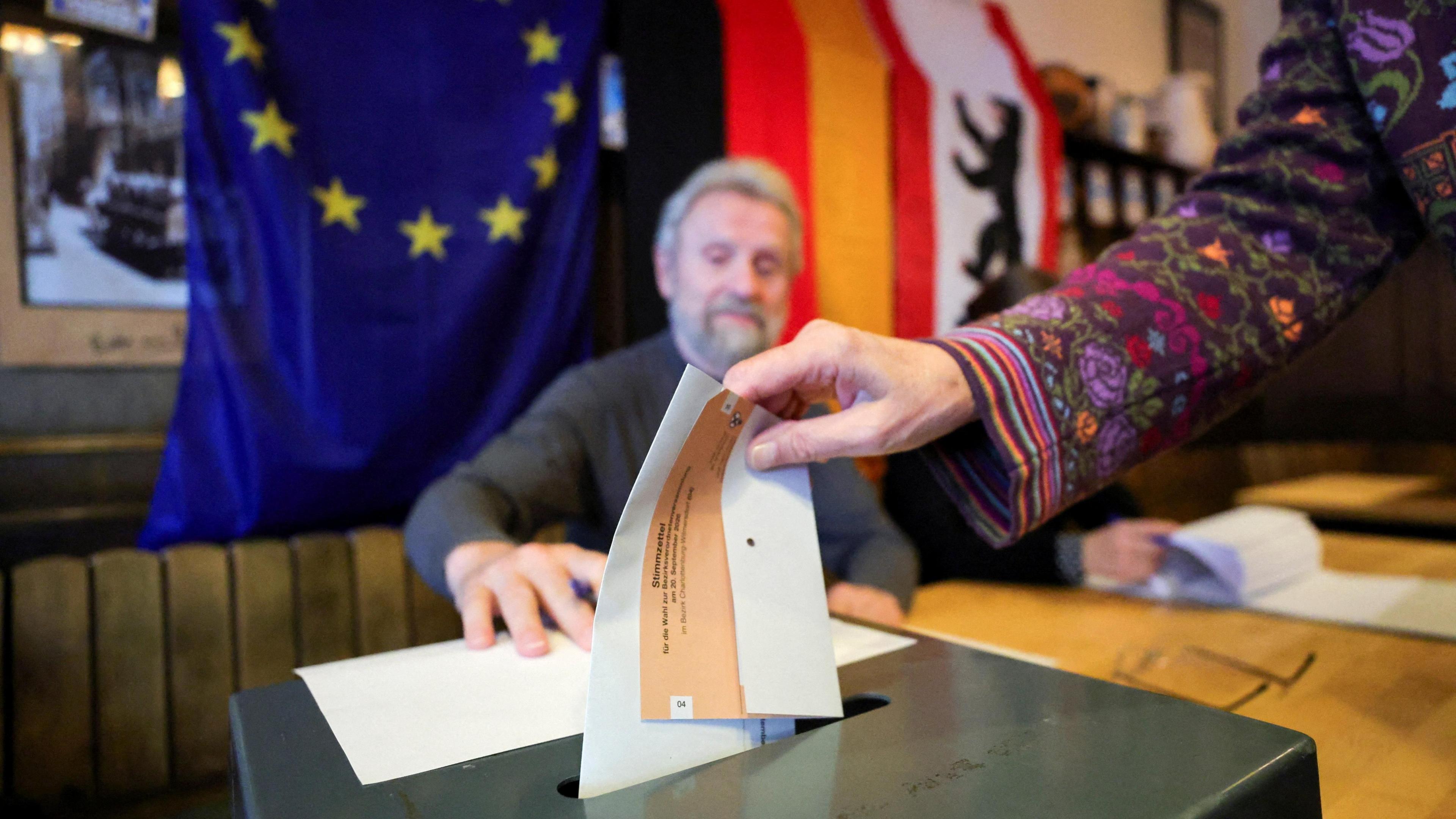 A close-up shot of a woman placing her paper vote into a black ballot box. Only her hand is visible, and the sleeve of her jumper which is purple with multicoloured flowers. A man with white hair and a beard is sat behind the desk on which the ballot box sits, and watches her deposit the vote. Behind him are the EU, German and Berlin flags.