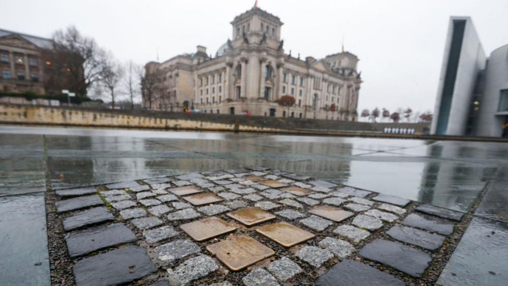 Ten brass plaques are placed among street stones on the floor in Berlin. In the background the Reichstag building can be seen.