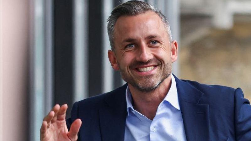 Ulrich Siegmund, who has greying hair and a short beard and wears a navy suit jacket with a light blue shirt, smiles and waves as he arrives at a press conference on Monday.