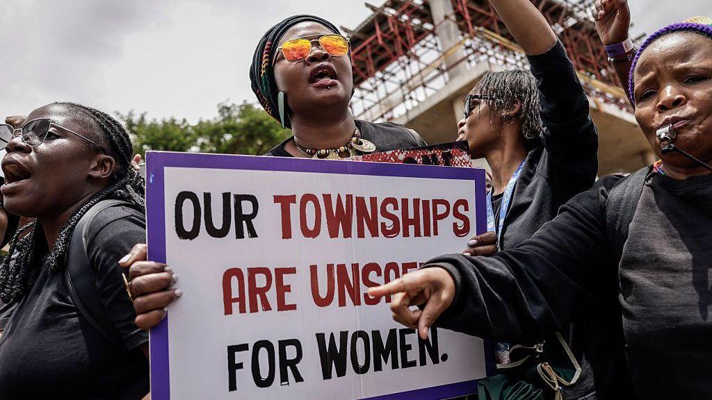 Women at a demonstration wearing black - one holds a placard that says: "Our townships are unsafe for women".