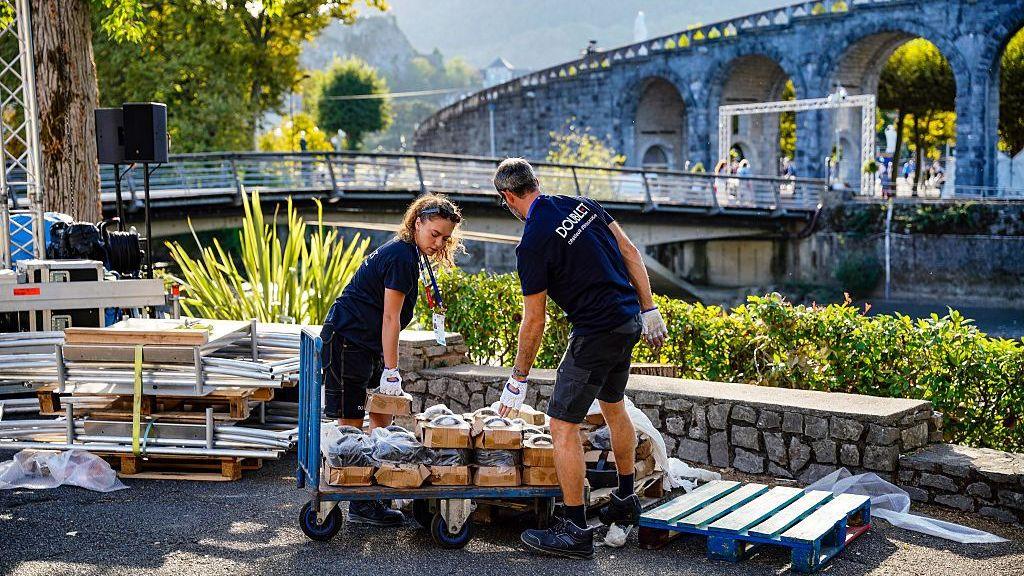 Two workers, a man and a woman dressed in navy polo shirts and shorts, move cardboard boxes off a trolley in the Sanctuary of Our Lady of Lourdes.