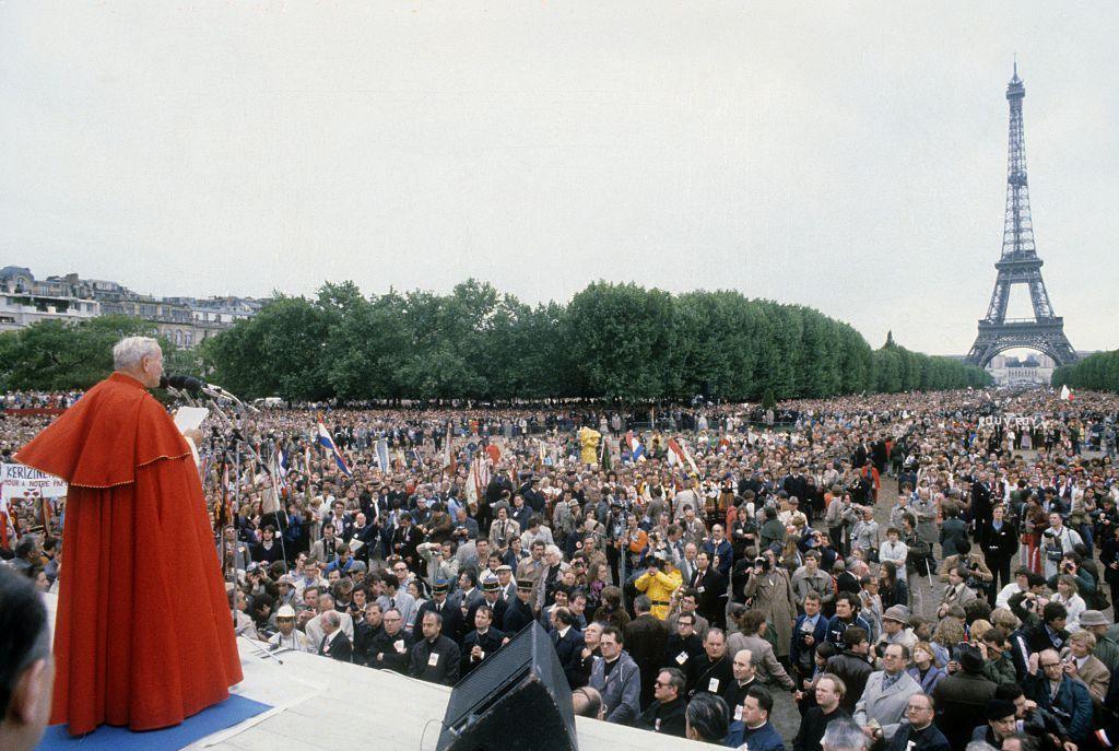 Pope John Paul II, wearing a red floor-length cape and pictured from the back, addresses a crowd of people in front of the Eiffel Tower in June 1980.