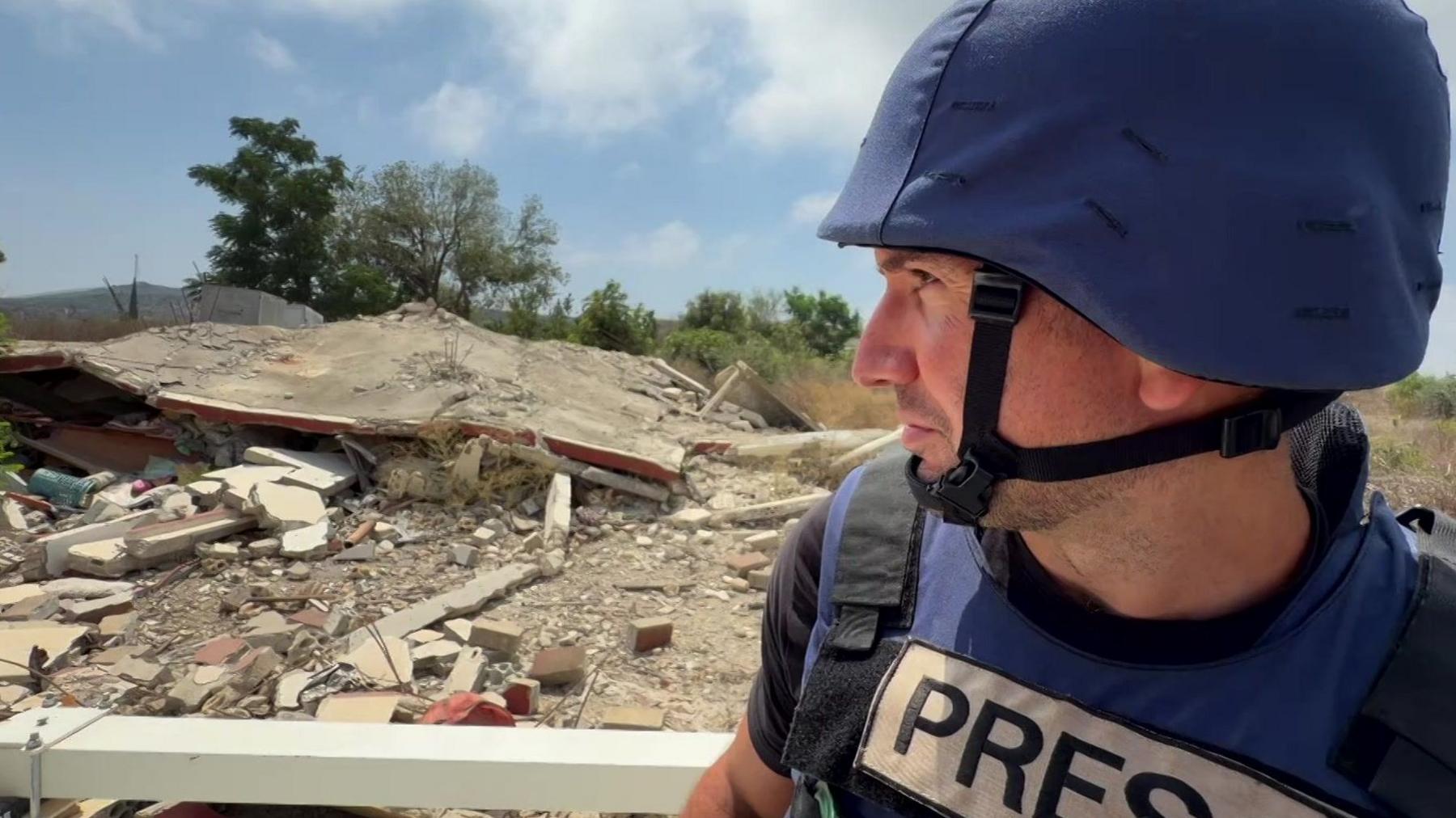 BBC correspondent Hugo Bachega, wearing a blue flak jacket and helmet, sits on top of a Unifil vehicle as it drives past destroyed buildings in southern Lebanon