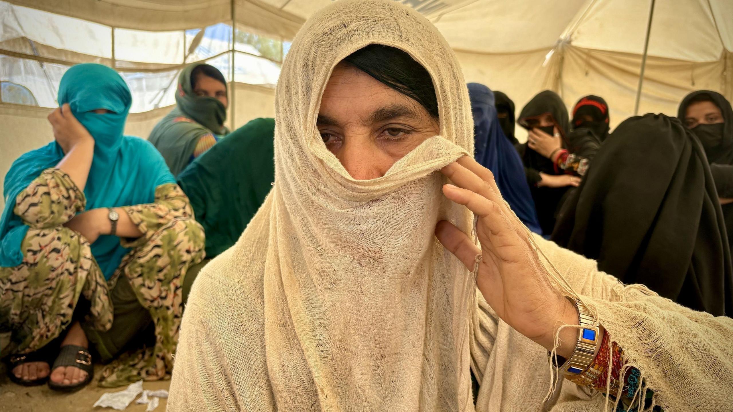 Zarina, an Afghan returnee, inside a tent with several others is using her veil to partially cover her face as she faces the camera.
