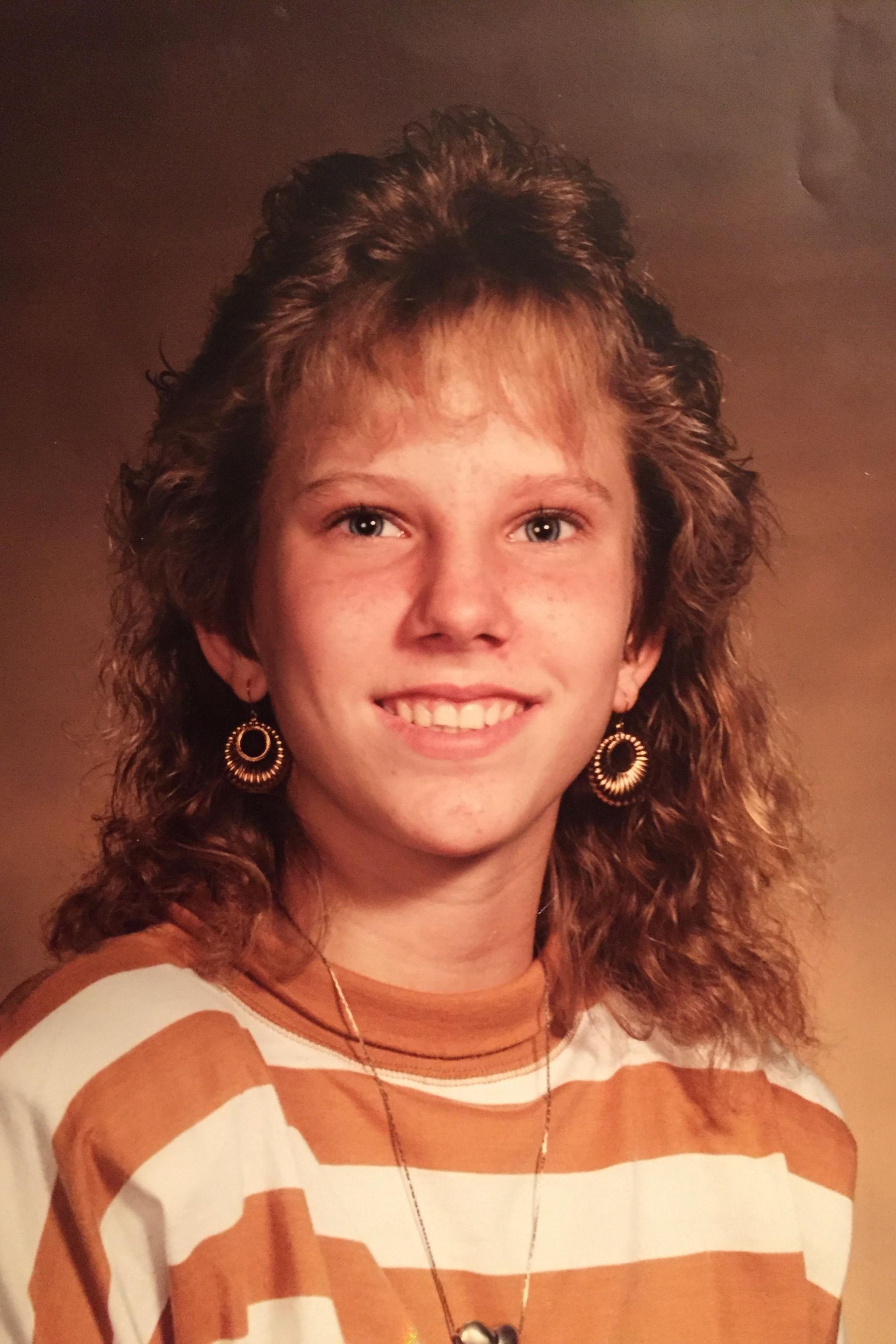 A portrait of Colleen Slemmer, taken in what looks like a photography studio with a backdrop. She is wearing a striped orange and white sweatshirt, gold drop earrings and is smiling at the camera. She has auburn hair and a fringe.